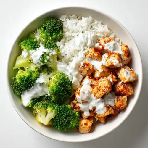 Overhead view of a High Protein Chicken Bowl Meal Prep featuring seasoned chicken pieces, white rice, and bright green broccoli, drizzled with white sauce.