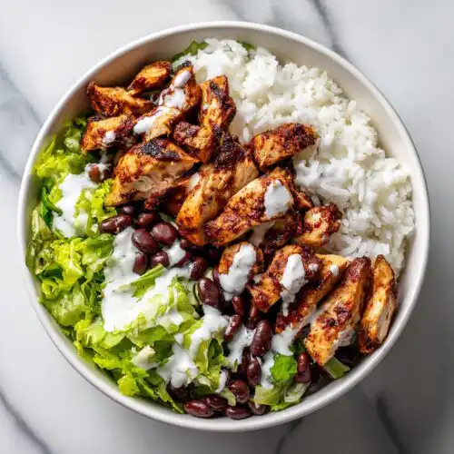 Overhead view of a High Protein Chicken Taco Bowl featuring seasoned chicken, white rice, lettuce, black beans, and a white sauce.