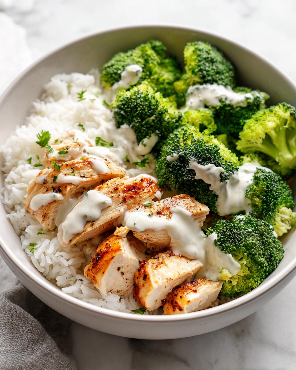 A bowl containing rice, sliced grilled chicken breast, and steamed broccoli drizzled with a creamy white sauce, perfect for High Protein Summer Meal Prep.