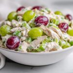 A white bowl filled with creamy Light Summer Chicken Salad mixed with celery and topped with red and green grapes.