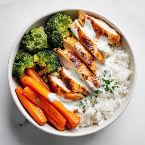 Overhead view of a Meal Prep Summer Dinner Bowls featuring sliced grilled chicken, white rice, roasted carrots, and broccoli, topped with a creamy sauce.