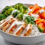 Close-up of a white bowl filled with rice, sliced grilled chicken, steamed broccoli, and carrots, drizzled with a light sauce, perfect for Meal Prep Summer Dinner Bowls.