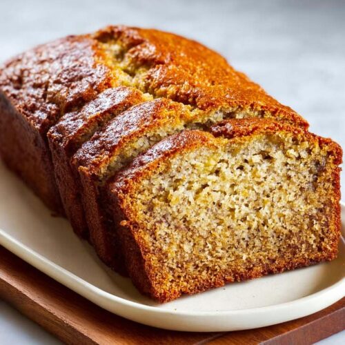 A freshly baked, golden-brown Moist Banana Bread Loaf, partially sliced on a white platter.