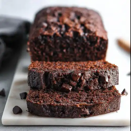 Two thick slices of moist Chocolate Banana Bread studded with chocolate chips, resting in front of the rest of the loaf.