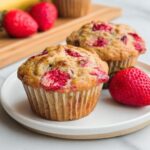 Two moist Strawberry Banana Muffins served on a white plate next to a fresh strawberry.