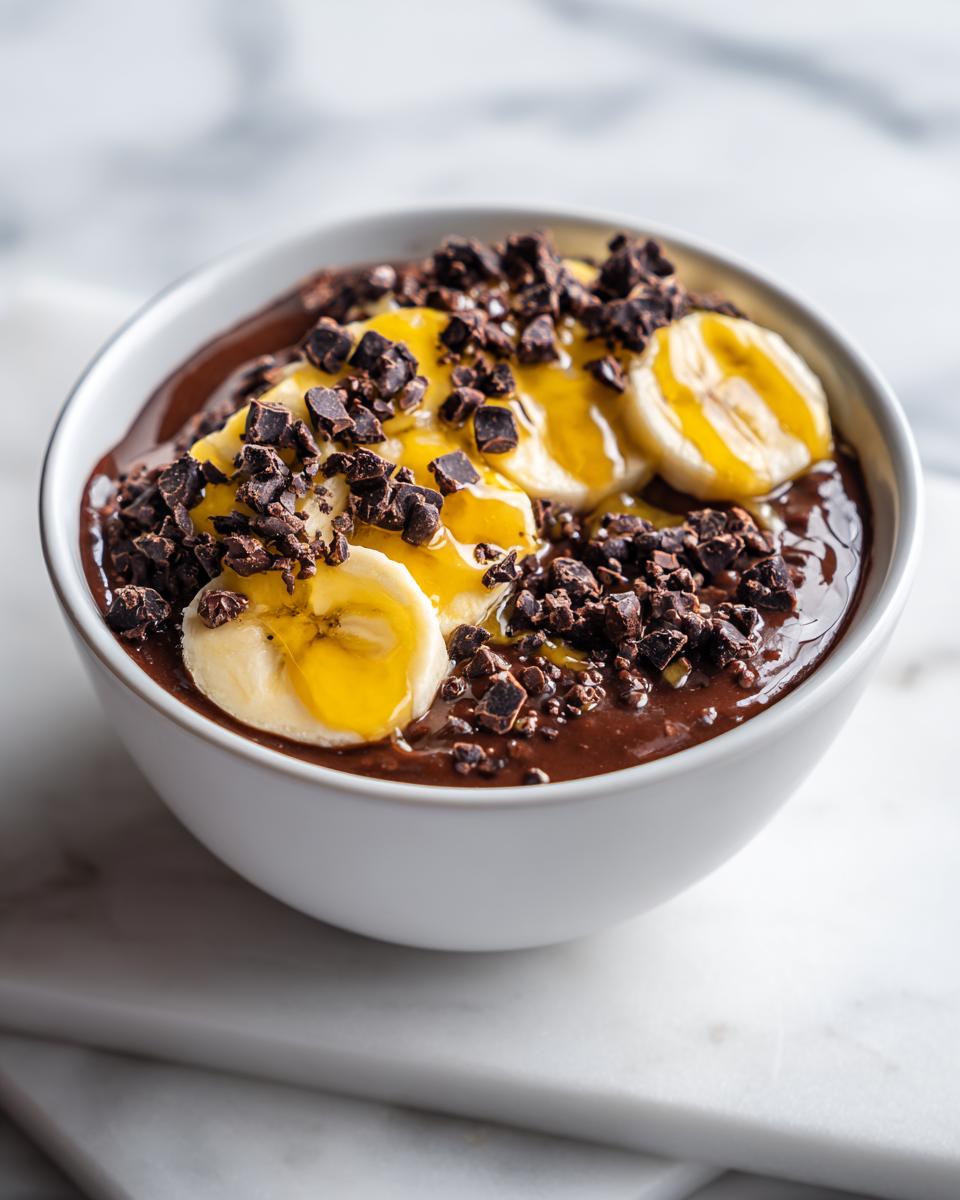 A white bowl filled with rich chocolate overnight oats, topped with sliced bananas, honey drizzle, and dark chocolate chunks.