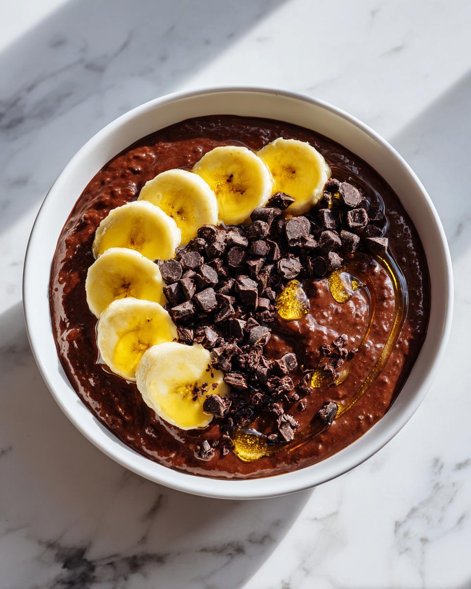 Overhead view of a bowl of rich Overnight Oats Chocolate topped with sliced bananas, chocolate chunks, and a drizzle of honey.