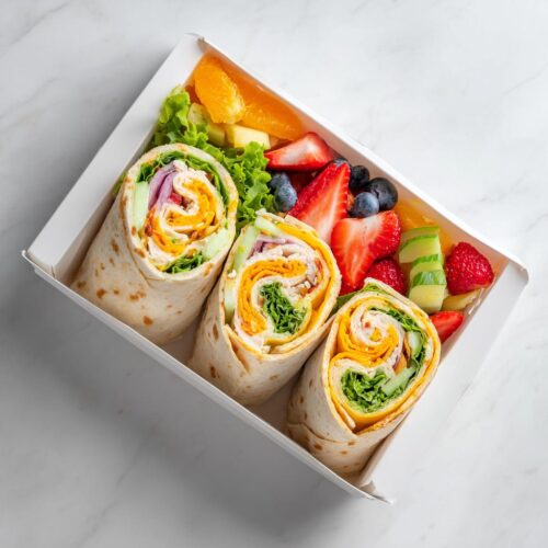 Three sliced Picnic Chicken Wrap Box rolls packed with lettuce, cheese, and meat, next to a fresh fruit salad.