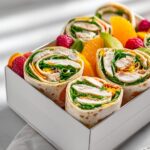 Close-up of sliced Picnic Chicken Wrap Box pinwheels filled with chicken, greens, and cheese, packed with orange slices and raspberries.