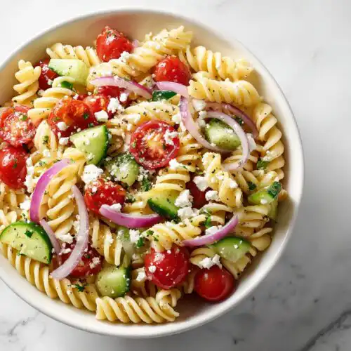 Close-up overhead view of a bowl of Picnic Pasta Salad featuring rotini pasta, cherry tomatoes, cucumber, red onion, and feta cheese.