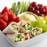A white dish filled with chicken wraps, strawberries, grapes, cucumber sticks, and hummus, perfect for Picnic Snack Box Ideas.