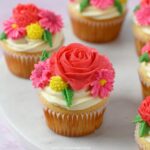 A close-up of a beautifully decorated Strawberry Wildflower Cupcake topped with red rose and pink daisy buttercream flowers.