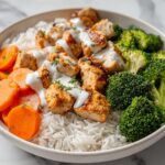 Close-up of a Summer Chicken and Rice Meal Prep bowl featuring grilled chicken, white rice, steamed broccoli, and sliced carrots with a white sauce.