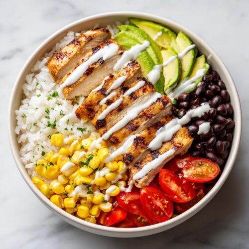 A vibrant Summer Chicken Burrito Bowl featuring grilled chicken, rice, corn, black beans, avocado, and tomatoes with a white drizzle.