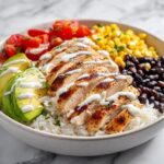 Close-up of a Summer Chicken Burrito Bowl featuring sliced grilled chicken, avocado, corn, black beans, and white sauce over rice.