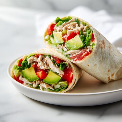Two halves of a fresh Summer Chicken Wrap Lunch showing shredded chicken, avocado, tomato, and lettuce.