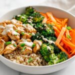 Close-up of a Summer Meal Prep Bowl featuring seasoned chicken, roasted broccoli, julienned carrots, and brown rice with a white sauce drizzle.