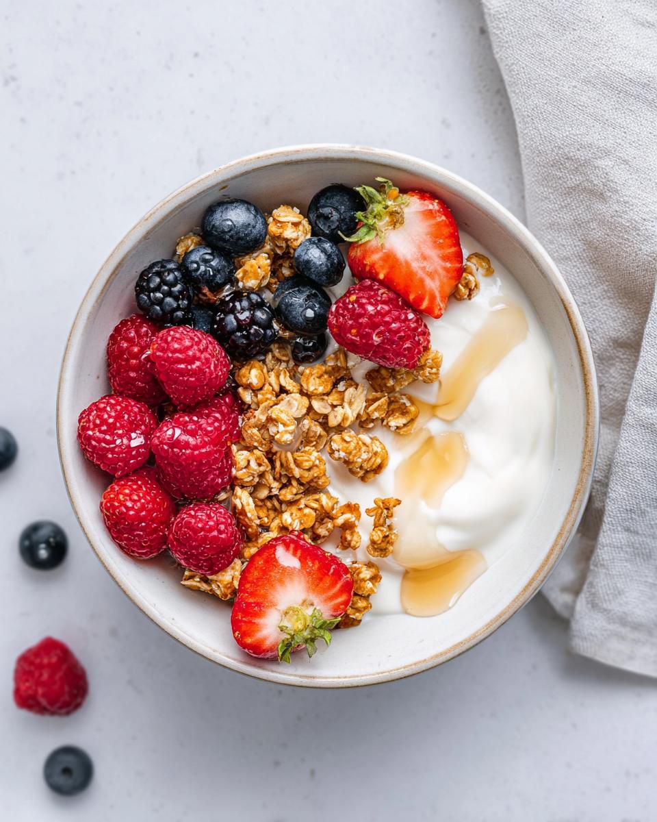 Overhead view of an aesthetic Yogurt Bowl with Fruits, featuring granola, raspberries, blueberries, strawberries, and a drizzle of honey.