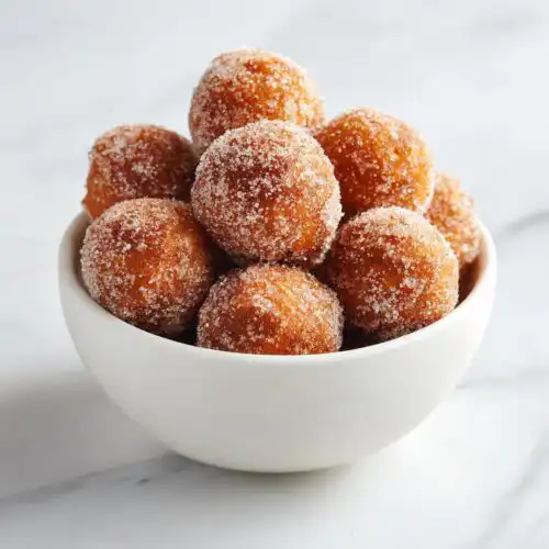 A white bowl filled with freshly made, golden brown Cinnamon Sugar Donut Bites coated heavily in sparkling sugar.