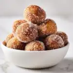 A white bowl overflowing with golden brown Cinnamon Sugar Donut Bites coated heavily in sparkling sugar.