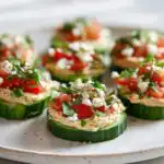 Several Mediterranean Cucumber Bites topped with hummus, diced tomatoes, feta, and herbs, arranged on a light plate.