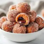 A close-up of sugar-coated Nutella Stuffed Churro Balls, with one broken open showing gooey melted chocolate filling.